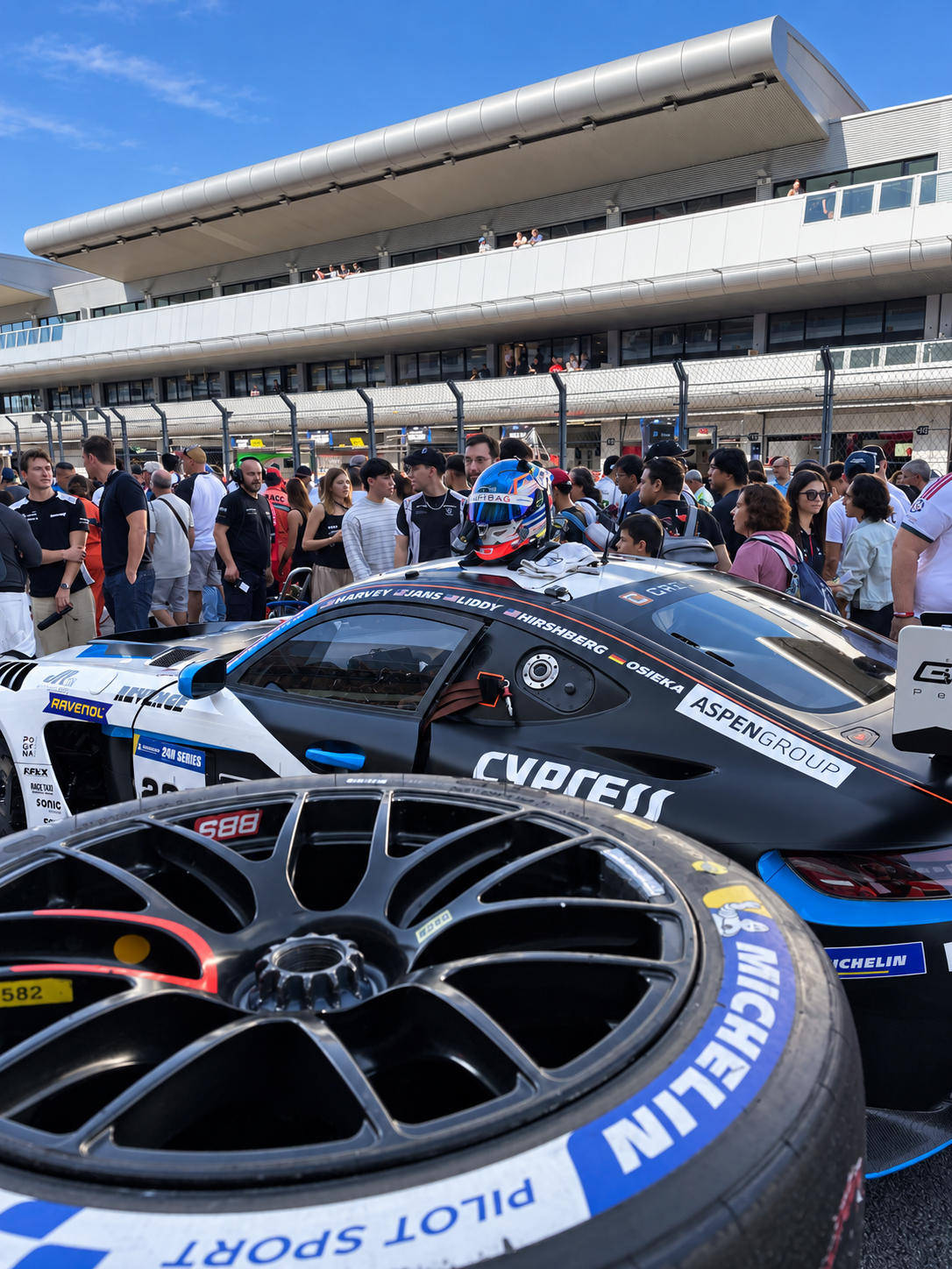 Rueda Michelin de competición en primer plano junto a un coche y público en la parrilla.