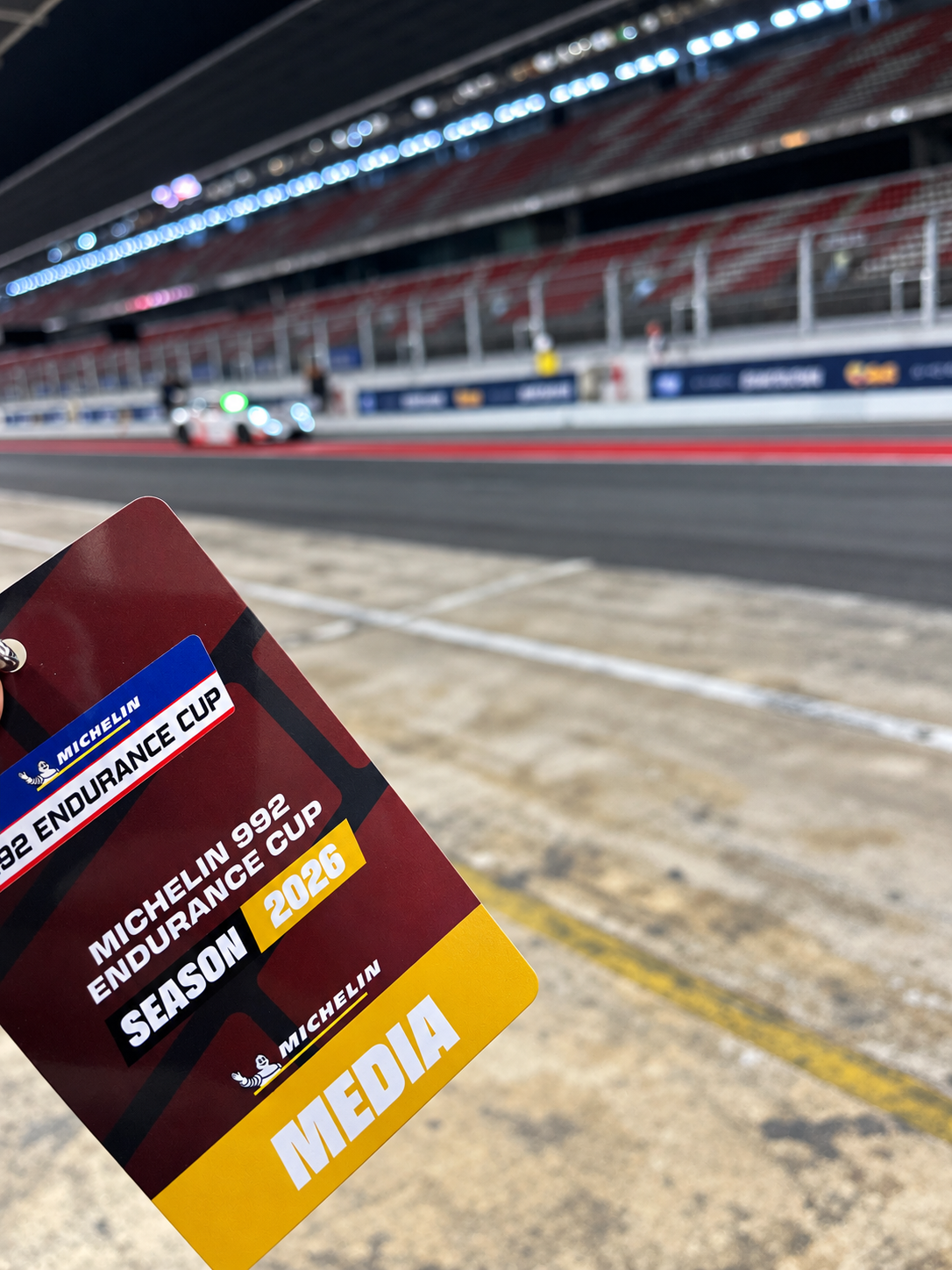 Acreditación de prensa en primer plano con el pit lane iluminado al fondo durante la noche.